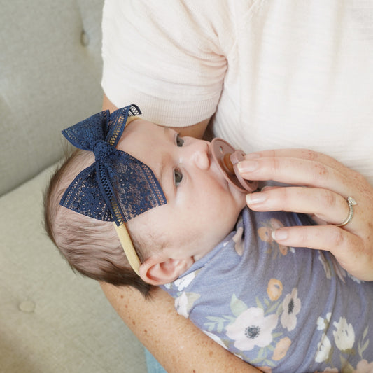 infant wearing Audrey navy blue lace bow, floral knit swaddle, and bibs pacifier village baby