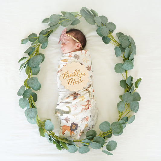 baby in wreath of greenery, wrapped in swaddle, with bow and custom name sign